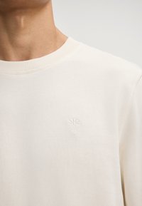 Kremowy sweter z okrągłym dekoltem i ściągaczami, z małym białym haftem kwiatu na klatce piersiowej. Miękka tekstura.