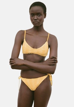 Ensemble de bikini au crochet jaune comprenant un bralette triangulaire avec de fines bretelles et une culotte nouée. La texture est en mailles ouvertes avec une finition lisse.