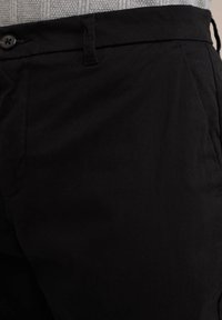 Pantaloni negri din bumbac cu croi drept, cu închidere cu nasture și două buzunare laterale. Textura vizibilă a țesăturii este netedă și mată.