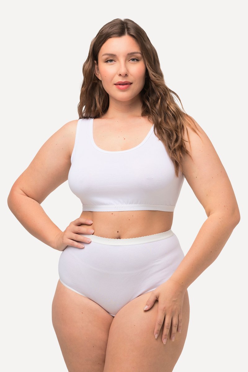 Plus-Size-Frau mit langen braunen Haaren, die ein weißes ärmelloses Croptop und hoch taillierte Slips vor einem einfarbigen Hintergrund trägt.