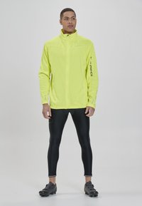 Helle gelbe, leichte Jacke mit hohem Kragen und Reißverschluss, figurbetontes Design, kombiniert mit schwarzen Leggings und eleganten schwarzen Sportschuhen.