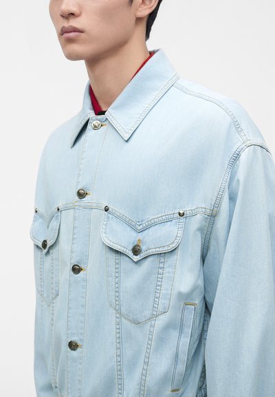Jeune personne portant une veste en jean bleu clair avec des boutons métalliques, des poches poitrine à rabat et des surpiqûres jaunes, montrée des épaules au menton.