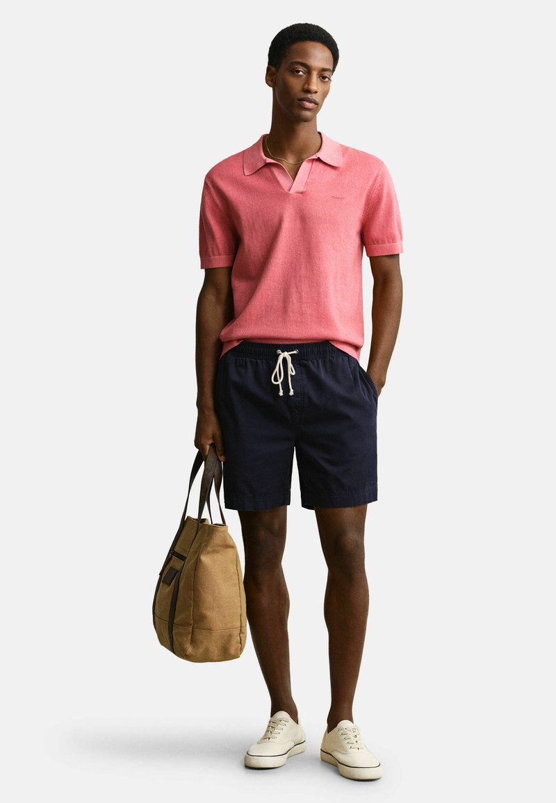 Un homme se tient debout, portant un polo à manches courtes rose, un short bleu marine avec cordon de serrage, des baskets blanches, et tenant un sac fourre-tout en toile beige à ses côtés.