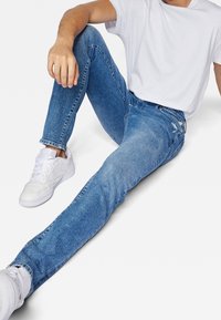 Blå denimjeans med slim fit och slitna detaljer, parat med vita sneakers, bärs med en åtsittande vit t-shirt, i sittande position.