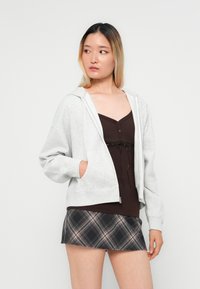 Svijetlo siva zip-up dukserica s prednjim džepovima, nosi se preko smeđeg topa bez rukava, uparenog s crnom kariranom mini suknjom. Glatka tekstura, ležeran kroj.