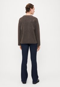 Brun strikket genser med avslappet passform og rillekant, parret med mørkeblå flagrende jeans og flate svarte sko. Utsikt fra baksiden.