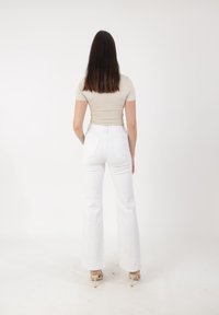 Mujer de pie de espaldas, vestida con una blusa ajustada de manga corta beige, pantalones blancos de tiro alto y sandalias de tacón beige.