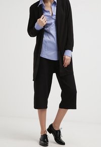 Cárdigan negro ligero sobre una camisa de botones azul claro, combinado con pantalones negros recortados y zapatos de cordones negros brillantes.