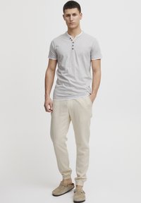 Camiseta gris de manga corta con tapeta de botones, combinada con pantalones jogger de color beige claro y zapatos grises sin cordones. Diseño simple y casual.