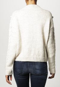 Kremowy sweter z dzianiny, z prążkowanym dekoltem i warkoczowymi zdobieniami na rękawach. Krótki krój i teksturowana tkanina.