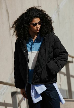 Frau mit lockigem Haar, die eine Sonnenbrille, eine schwarze gesteppte Jacke und ein blau-weißes Hemd trägt, steht draußen vor einer hellen Wand.