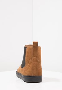 Brązowe zamszowe botki Chelsea z czarnymi elastycznymi panelami bocznymi i pętelką. Posiadają płaską czarną gumową podeszwę oraz gładką teksturę.