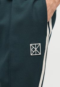 Zbliżenie na ciemnozielone spodnie z białymi paskami po bokach oraz geometrycznym, kwadratowym logo w pobliżu kieszeni, ręka częściowo włożona do kieszeni.