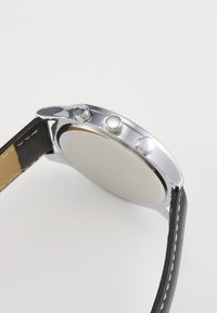 Boîtier de montre en métal argenté avec un cadran rond et lisse. Bracelet en cuir noir avec des coutures blanches. Trois poussoirs décoratifs sur le côté.