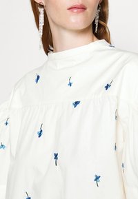 White cotton blouse featuring blue floral embroidery, with a round neckline and puffed sleeves. Includes a gathered seam for added texture.