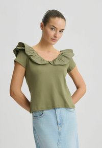 Oliven grøn top med en flæsekrave og korte ærmer, lavet af blødt stof. Båret med en lys blå denimnederdel.