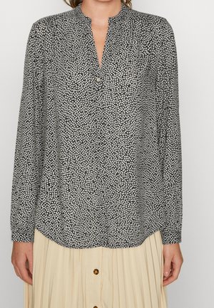 Femme portant un chemisier à manches longues à pois noirs et blancs avec un col rond et une jupe plissée beige avec des boutons.
