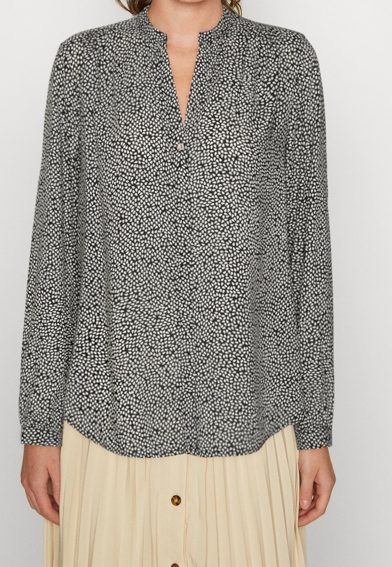 Femme portant un chemisier à manches longues à pois noirs et blancs avec un col rond et une jupe plissée beige avec des boutons.