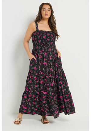 Kobieta w czarnej maxi sukience w różowy kwiatowy wzór, z szerokimi ramiączkami, marszczonym gorsetem, kieszeniami i złotymi sandałami, stojąca na jednolitym tle.