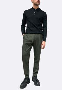 Polo nera a maniche lunghe con abbottonatura a bottoni, abbinata a pantaloni verdi scuro a pieghe e sneakers nere. Design semplice, texture liscia.