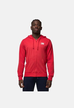 Uomo in piedi che indossa una felpa con cappuccio rossa con zip e logo "LEONE 1947" sul petto e pantaloni scuri su uno sfondo bianco semplice.