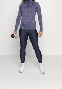 Granatowe legginsy i teksturowana fioletowa bluzka z długim rękawem i zamkiem przy kołnierzu, uzupełnione białymi trampkami i butelką wody w ręku.