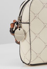 Sac en cuir à texture crème avec un motif en diamant, garniture noire et ferrures en métal. Présente une étiquette métallique avec le nom de la marque attachée.