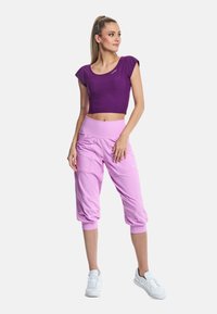 Violetinis trumpas viršus derinamas su rožinėmis capri kelnėmis, turinčiomis susiraukšlėjimo akcentus ir elastingą juosmenį. Modelis dėvi baltus sportbačius.