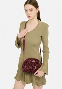 Bordeauxfarbene, gesteppten Handtasche mit geschwungener Form, goldenem Logo und einem dünnen, verstellbaren Trageriemen, kombiniert mit einem gerippten, olivgrünen Kleid.