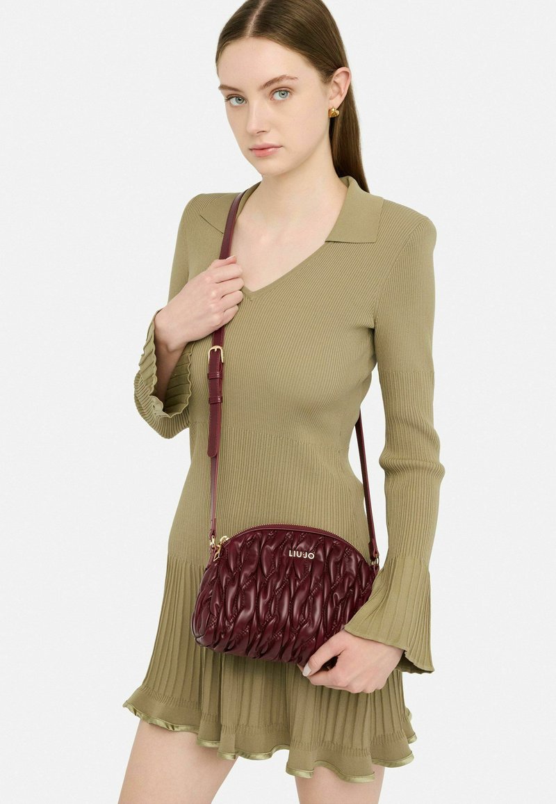 Bordeauxfarbene, gesteppten Handtasche mit geschwungener Form, goldenem Logo und einem dünnen, verstellbaren Trageriemen, kombiniert mit einem gerippten, olivgrünen Kleid.