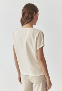 Blusa a maniche corte beige chiaro in un tessuto morbido e liscia. Presenta un collo rotondo e una vestibilità rilassata, vista da dietro.