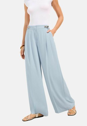 Femme portant un pantalon large taille haute bleu clair avec des boutons au niveau de la taille, un haut blanc sans manches et des sandales beige.