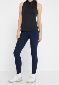 Černé sleeveless polo tričko s knoflíkovou légou, kombinované s námořnicky modrými legínami s bočními panely a bílými sportovními botami.