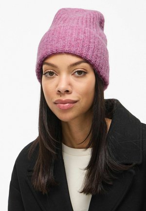 Bonnet en tricot rose avec un ajustement décontracté, motif texturé et matériau en laine douce, porté avec un manteau noir et un haut de couleur claire.