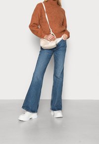 Femme portant un pull en maille couleur rouille, un jean large bleu, des baskets écourtées blanches et portant un sac à bandoulière beige, se tenant devant un mur blanc.