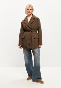Chaqueta de plumas marrón con cintura ceñida, cuello con muesca y bolsillo de parche, combinada con unos vaqueros acampanados de color azul claro y zapatillas de ante marrón.