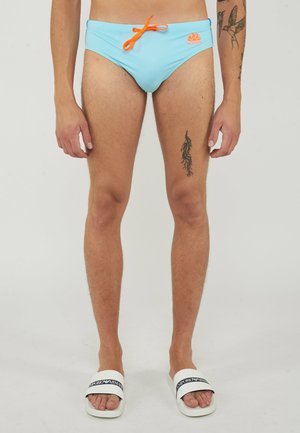 Uomo che indossa slip da bagno azzurro chiaro con coulisse arancione e logo, in piedi con tatuaggi visibili su gambe e braccia, ciabatte bianche ai piedi.