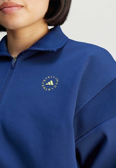 Sudadera azul marino con cremallera y cuello alto, con un logotipo circular en color amarillo claro. Tejido suave con acentos texturizados en las mangas.