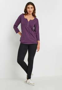 Lila långärmad topp med V-ringning och knappar, prydd med subtila veck längs fronten. Kombinerad med svarta slim-fit jeans och vita sneakers.