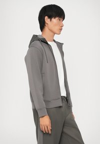 Felpa grigia con zip e cappuccio foderato in grigio scuro, caratterizzata da una vestibilità rilassata, maniche lunghe e tasche frontali, indossata sopra una maglietta bianca.