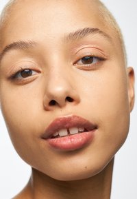 Gros plan d'un visage avec une peau pâle, des cheveux blonds courts, de légers grains de beauté, des yeux marron et des lèvres hydratées d'une teinte rose naturel.