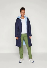 Ναυτικό αδιάβροχο παλτό με ανοιχτό μπλε κουκούλα, που διαθέτει πλαϊνές τσέπες, μεσαίου μήκους σχεδίαση και φοριέται πάνω από λευκό μπλουζάκι και πράσινα παντελόνια.