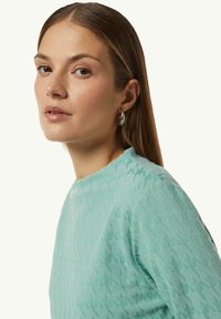 Hellblau strukturierter Pullover mit rundem Ausschnitt und gemusterten Details; kombiniert mit silbernen Ohrhängern.