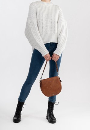 Personne portant un pull en tricot blanc, un jean skinny bleu, des bottes noires à lacets, tenant un sac à main en daim marron avec une bandoulière tressée.