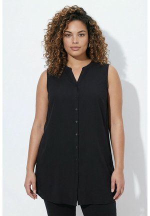 Kvinde med krøllet hår og fregner iført en ærmeløs sort tunika med knapper på en ensfarvet lys baggrund.