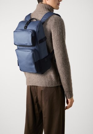 Zaino blu con due tasche frontali, realizzato in materiale resistente, dotato di spallacci regolabili e un design testurizzato, indossato sopra un maglione beige.