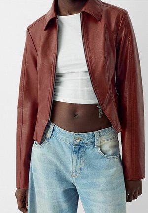 Osoba o ciemnej karnacji ubrana w otwartą brązową skórzaną kurtkę, biały prążkowany krótki top i jasnoniebieskie dżinsy.