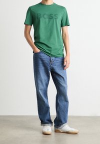 Vihreä puuvillapaita, jossa on "BOSS"-logo, yhdistettynä sinisiin farkkuihin. Valkoiset lenkkarit, joissa on kumipohjat, neutraali tausta.