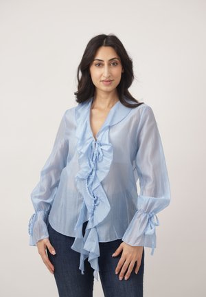 Frau trägt eine durchsichtige hellblaue Bluse mit Rüschendetails vorne und gebundenen Manschetten, steht vor einem schlichten hellen Hintergrund.