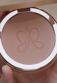 Compact bronzer met een zachte, matte, lichtbruine poeder. Bevat een reliëfontwerp van een vlinder, verpakt in een ronde, metallic doos.